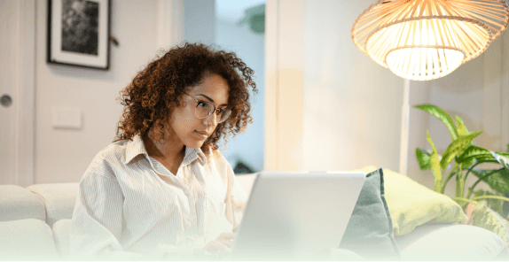 Person sitting on a couch using a laptop in a cozy living room with a hanging lamp and green plant nearby.