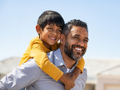 Child riding on an adult’s back outdoors with a clear blue sky and rooftops in the background.