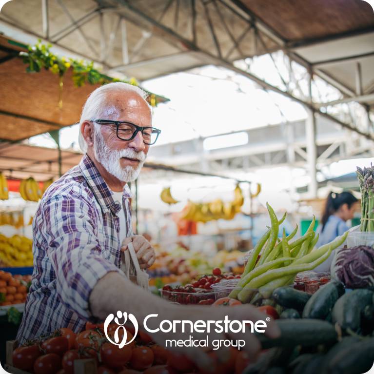 Person selecting fresh vegetables at an outdoor market with Cornerstone Medical Group logo at the bottom.